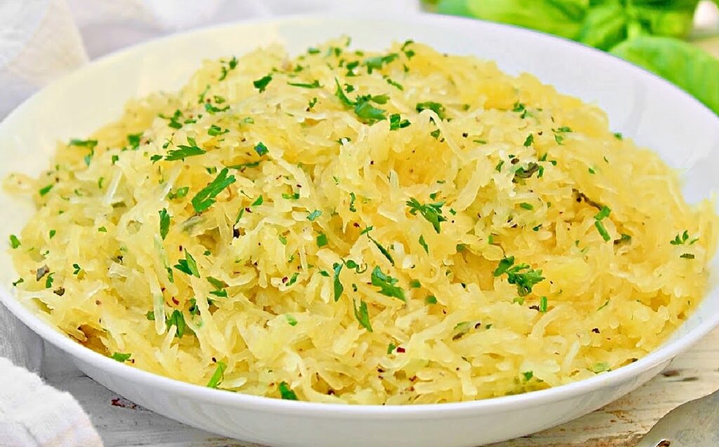 Side view of white bowl filled with cooked spaghetti squash. Fresh green herbs are sprinkled over the top. Green herbs are in the background.